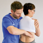 Therapeut stabilisiert den Schultergürtel der sitzenden Patientin und schiebt mit der rechten Hand den Angulus costae nach ventral