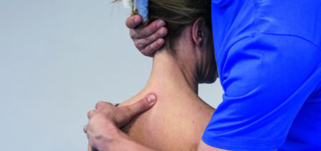Nahaufnahme von hinten: Therapeut stabilisiert den nach rechts gedrehten Kopf der Patientin und legt die andere Hand an die Halswirbelsäule
